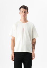 Male model stands against a white background, wearing a cream t-shirt with a pink rose graphic, paired with black pants.