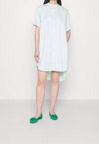 Robe verte claire à boutons, avec des manches courtes volantes, un design fluide et des fentes sur les côtés. Portée avec des chaussures plates vertes ornées d'un accent décoratif.