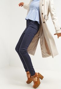 Marina blå skinny jeans kombinerat med en ljusblå peplumtop och en krämfärgad trenchcoat, kompletterat med tan plattformsskor med en kraftig klack.