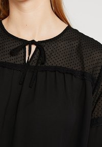 Schwarze Bluse mit transparentem gepunktetem Einsatz, Spitzenbesatz und rundem Ausschnitt mit einer Bindeschleife vorne. Weicher, fließender Stoff.