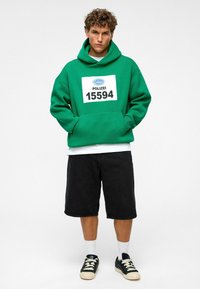 Groene hoodie met een grote witte "POLIZEI 15594" opdruk, gecombineerd met zwarte shorts en zwarte sneakers met crèmekleurige accenten.