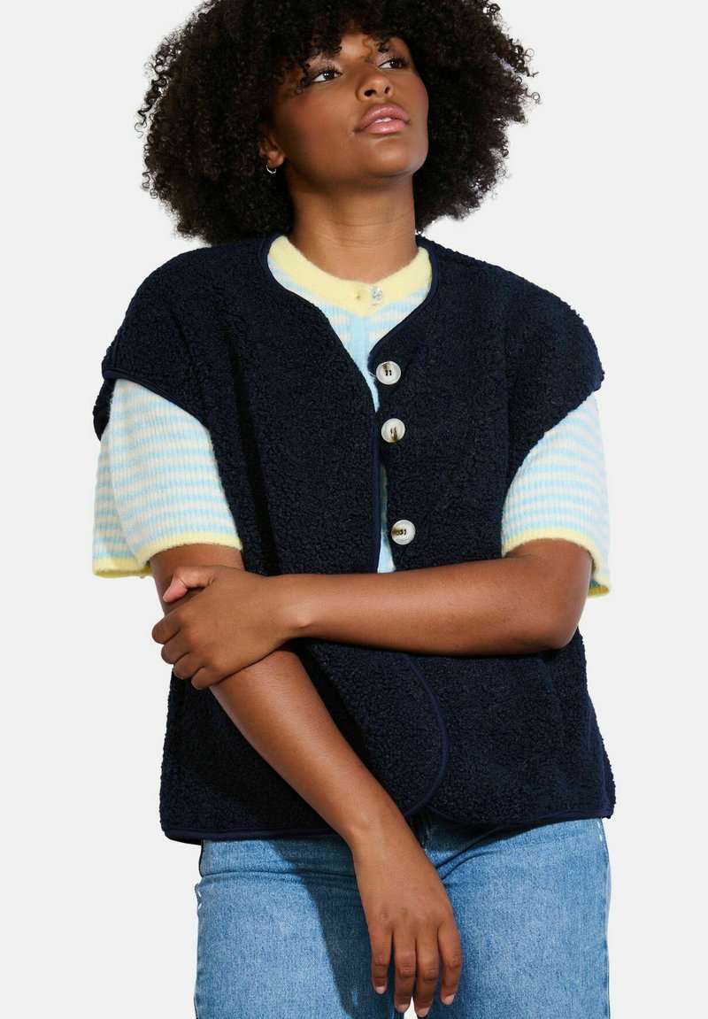 Gilet in pile blu marina con chiusura a bottoni, orlo arrotondato e maniche corte, indossato sopra una camicia a righe azzurro chiaro e gialla, abbinato a pantaloni in denim.