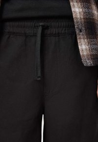Pantalons noirs avec une taille élastique et un cordon de serrage. Le tissu est lisse, avec une poche latérale. Accentué par une chemise à carreaux marron.