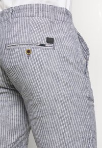 Pruhované kalhoty z lehkého materiálu, které mají zadní kapsu na knoflík a označení značky. Barvy jsou modrá a bílá s jemným pruhovaným vzorem.