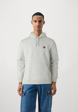 Sudadera gris clara con cordón, bolsillo frontal y un pequeño logo multicolor en el pecho. Combinada con jeans de cargo azules.