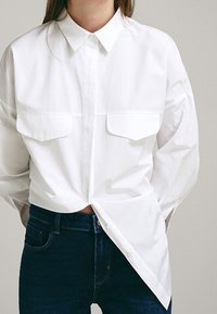 Camicia bianca con bottone, dotata di colletto classico, due tasche sul petto e vestibilità rilassata. Tessuto morbido e testurizzato abbinato a denim scuro.