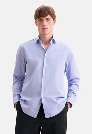 Jeune homme aux cheveux châtain clair porte une chemise bleu clair à boutons et un pantalon noir, debout les mains dans les poches devant un fond blanc.