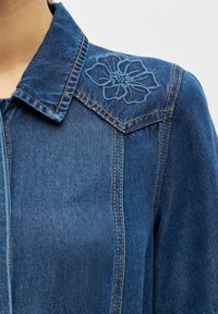 Détail de l'épaule et du col d'une veste en denim avec des surpiqûres contrastantes et un motif floral brodé sur la partie supérieure de l'épaule.