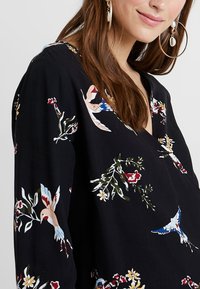 Femme portant un haut noir avec un imprimé de oiseaux colorés et de fleurs, des boucles d'oreilles en or, et de longs cheveux bruns sur les épaules.