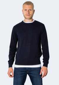 Maglione a girocollo blu navy con maniche lunghe, realizzato in tessuto morbido. Presenta un logo sottile sul petto e una vita aderente. Indossato sopra una maglietta bianca.