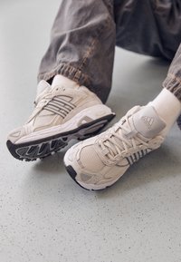 Bílé síťované sportovní tenisky s šedými akcenty a texturovanými podrážkami. Obsahují logo na zadní straně, tkaničky a ventilační otvory.