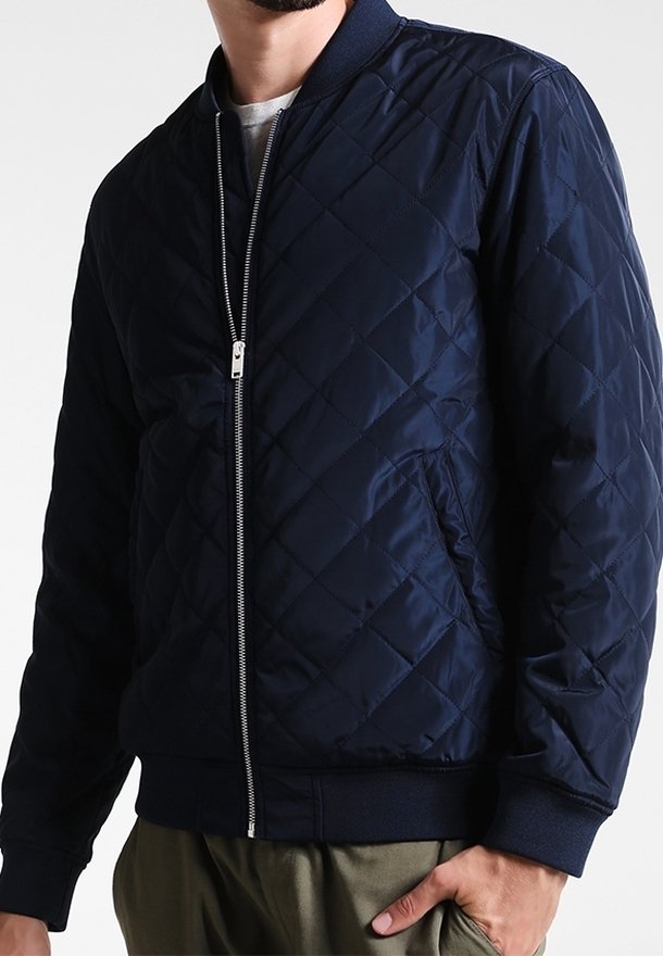 Marinblå quiltad bomberjacka med dragkedja framtill, ribbade ärmar och krage. Har sidofickor och en slät, glänsande yta.