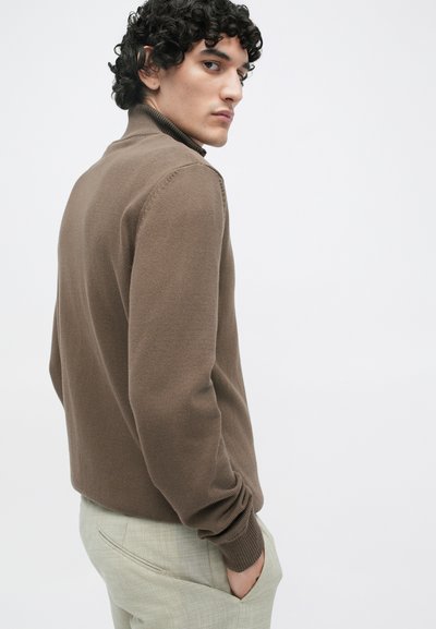 Pull en tricot marron avec un col montant, des poignets côtelés et un motif subtilement texturé, associé à un pantalon texturé de couleur claire.