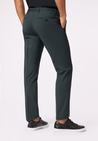 Pantaloni su misura di un verde scuro con vestibilità slim, caratterizzati da una texture liscia, tasche posteriori e chiusure a loop, abbinati a sneaker nere.