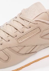 Gros plan sur une basket Reebok beige montrant des lacets texturés, des détails en cuir cousu, un logo embossé et une semelle extérieure en caoutchouc marron clair.