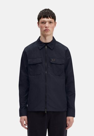 Giacca zip-up navy con due tasche sul petto, colletto ribassato e branding discreto. Texture liscia e materiale leggero.