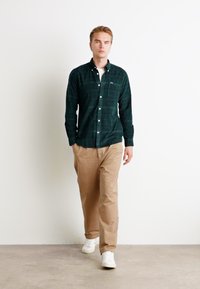 Camisa de flanela xadrez verde escuro, abotoada, com uma t-shirt branca por baixo. Calças bege e ténis brancos completam o conjunto. Fundo liso.