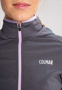 Mujer que lleva una chaqueta gris con cremallera y detalles en lavanda, con el logo "COLMAR" en el pecho, mostrando solo la parte inferior de la cara y la parte superior del torso.