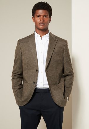 Mann trägt einen braunen Tweed-Blazer über einem weißen Hemd mit Knöpfen und dunklen Hosen, steht mit Händen in den Taschen vor neutralem Hintergrund.