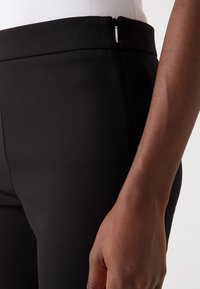 Černé přiléhavé kalhoty s hladkým materiálem, minimalistickým designem a zipem v oblasti pasu. Detailní záběr ukazující strukturu a umístění ruky.