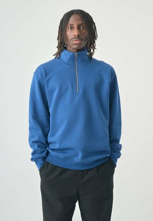 Blauer Fleece-Pullover mit hohem Kragen und halbem Reißverschluss, ausgestattet mit langen Ärmeln und einer lockeren Passform. Weiche Textur und minimalistisches Design.