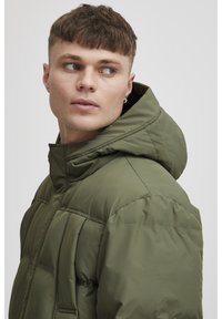 Olive green puffer jacket with a hood, featuring a quilted design, zippered front, and visible stitching. Lightweight fabric with a smooth texture.