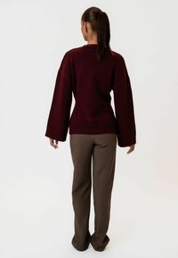 Burgundski oversized pulover s širokim rukavima, pletenom teksturom i rebrastim manžetama, u kombinaciji s smešnim odrezanim hlačama, viđen sa leđa.