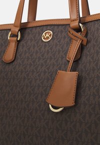 Brun mønstrede tote taske med beige læderhåndtag, guldfarvet hardware og et vedhæng. Logoet er præget i midten af tasken.