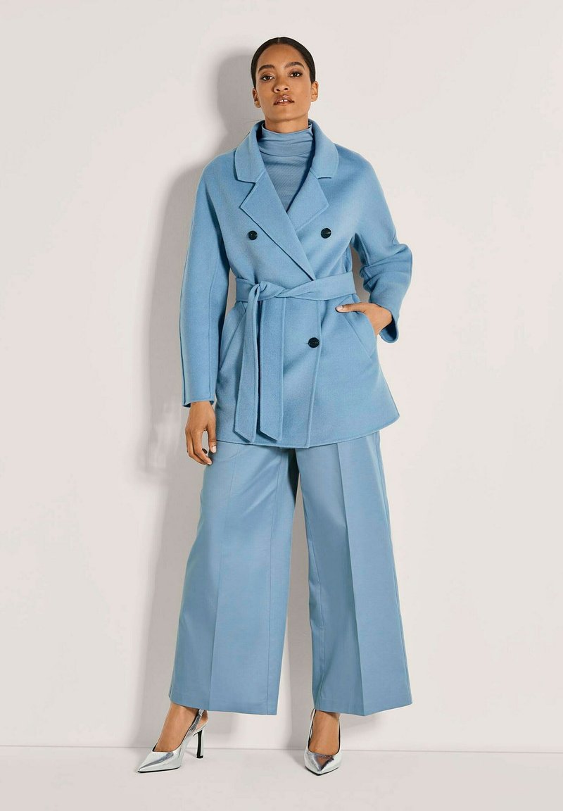 Vrouw in een lichtblauwe, met een riem afgewerkte, dubbelgeknoopte jas, met wijde broekspijpen en zilveren puntige hoge hakken, staande tegen een eenvoudige muur.