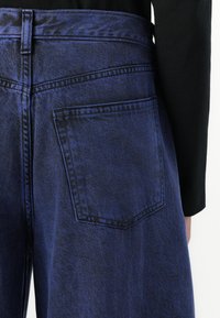 Faded blue denim jeans with a high waistband, featuring a single back pocket. Smooth texture and contrasting stitching detail.