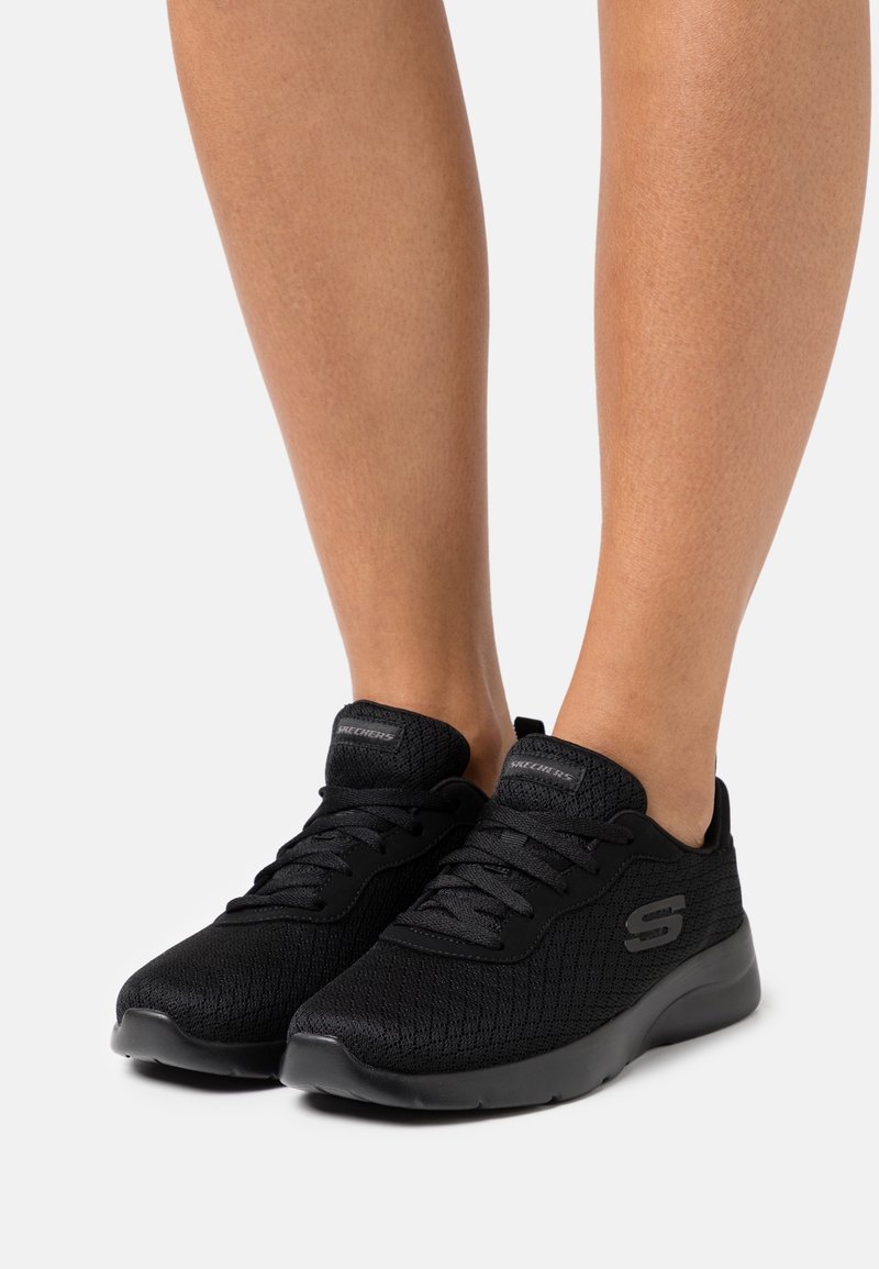 Personne portant des baskets Skechers noires en mesh à lacets avec semelles rembourrées, debout sur un fond clair uni.