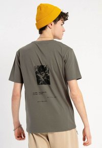 Oliwkowa bawełniana koszulka z krótkim rękawem z czarnym nadrukiem graficznym na plecach, ozdobiona abstrakcyjnymi wzorami i szczegółami tekstowymi. Luźny krój.