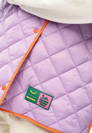 Fioletowa pikowana tkanina z pomarańczową lamówką i zatrzaskami, z zielono-różową naszywką z napisem „Mango Explorers” oraz grafiką gór.