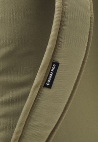 Primo piano di una cinghia di zaino in tessuto khaki che mostra un'etichetta del brand CONVERSE in bianco e nero.