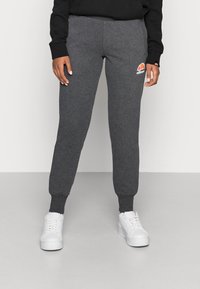 Persoon in donkergrijze joggers met een ellesse-logo, zwart lang mouwen top en witte platform sneakers staat op een witte vloer.