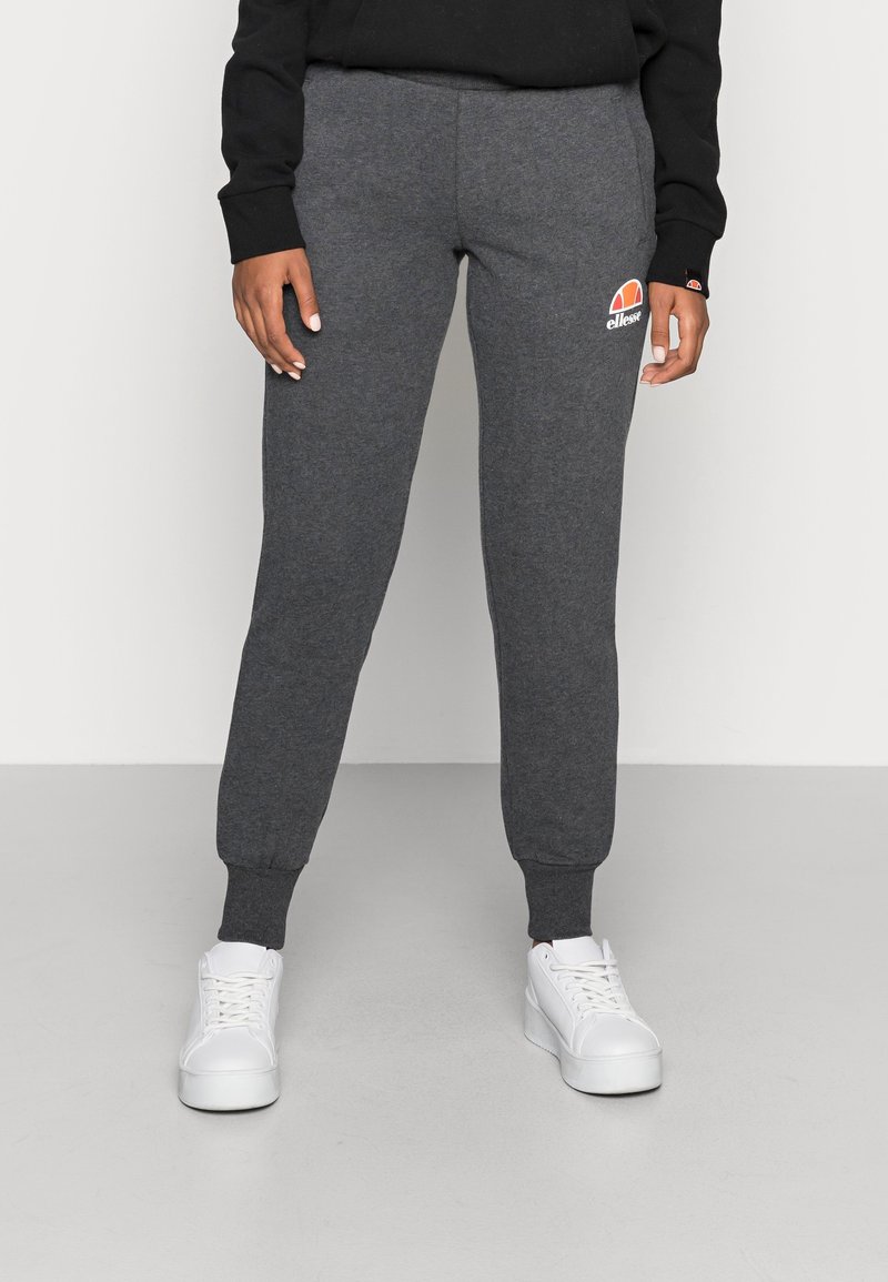 Persoon in donkergrijze joggers met een ellesse-logo, zwart lang mouwen top en witte platform sneakers staat op een witte vloer.