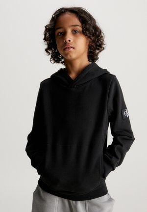 Sweatshirt preta em tecido macio com um bolso canguru, punhos e barra canelados, apresentando um patch de logo na manga esquerda. Textura suave.