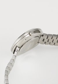 Montre en acier inoxydable argenté avec un boîtier rond lisse et un bracelet à maillons. Présente un logo sur le côté et un fond de montre texturé.