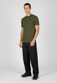 Olivegrünes kurzärmliges T-Shirt mit Rundhalsausschnitt und kleinem schwarzem Logo auf der Brust, kombiniert mit lockeren schwarzen Hosen und schwarzen Sneakers.