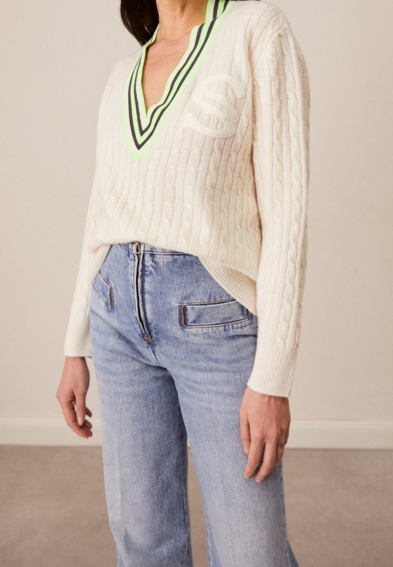 Cremefarbener Kabelstrickpullover mit tiefem V-Ausschnitt und grünem sowie schwarzem gestreiftem Kragen. Kombiniert mit hoch taillierten hellblauen Jeans.