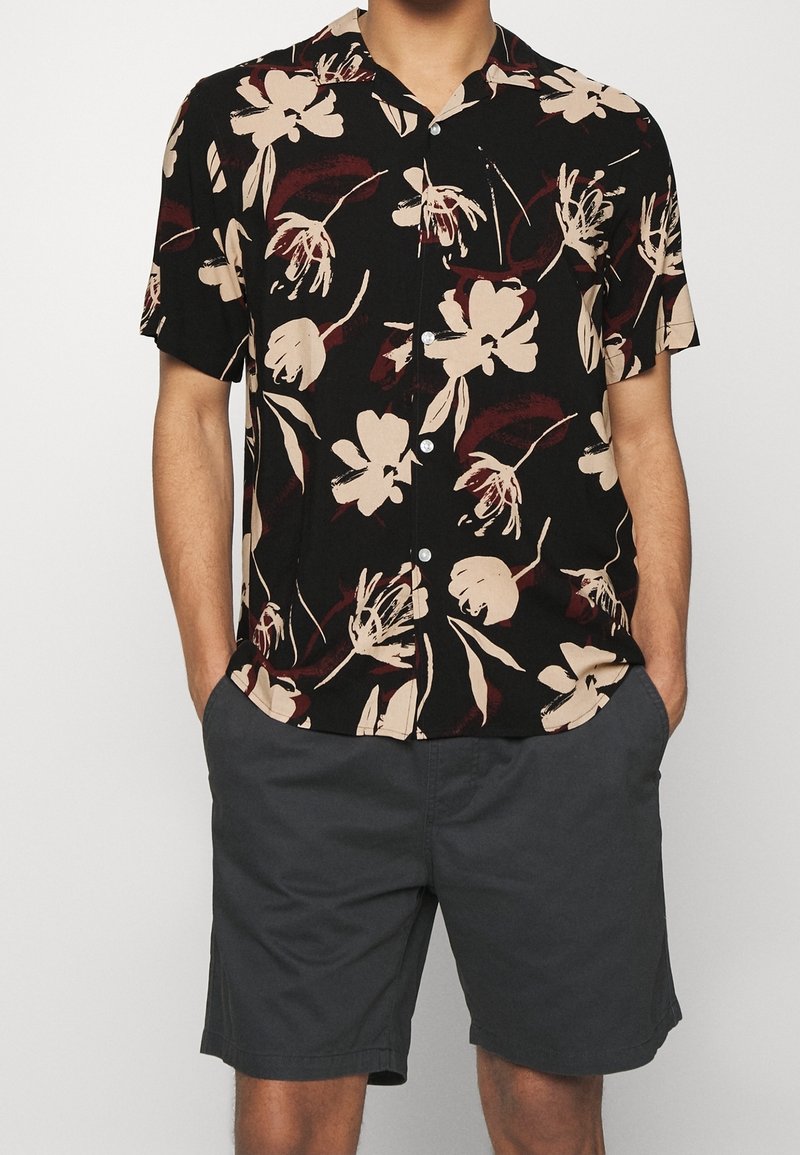 Lyhythihainen musta paita, jossa on kermaan ja viininpunaan vivahtava kukkakuosi, yhdistettynä harmaisiin shortseihin. Materiaali vaikuttaa kevyeltä ja istuvuus rento.