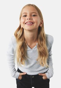 Langærmet grå henley skjorte med struktureret stof, to knapper ved kraven, parret med mørke jeans. Barnet smiler med hænderne i lommen.