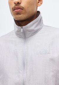 Gros plan d'une personne portant une veste zippée gris clair avec un col haut et un logo discret sur la poitrine.