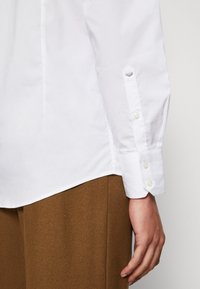 Camisa branca de botão com uma textura suave, apresentando um colarinho pontudo, mangas longas e botões semelhantes a pérolas. Calças de tecido castanho parcialmente visíveis.