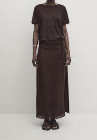 Femme portant un haut marron foncé à manches courtes et une jupe longue assortie avec une fente latérale, des sandales noires et un bracelet argenté au poignet droit.