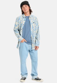 Blå mönstrad skjorta med knappar över en blå t-shirt, tillsammans med ljusa jeans och svarta sneakers. Skjortan har abstrakta krämfärgade mönster.