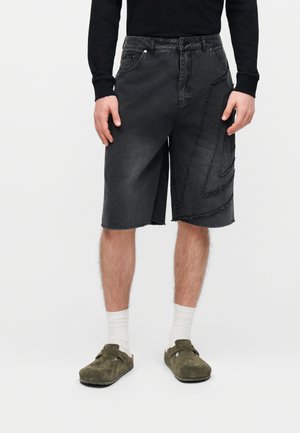 Hombre con camiseta negra de manga larga, pantalones cortos de mezclilla negros hasta la rodilla con patrones desgastados, calcetines blancos y zapatos slip-on verde oliva.