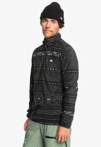 Svart fleece-pullover med geometriska mönster i grått och grönt. Har en hög krage och en dragkedja fram. Bärs med gröna cargobyxor.