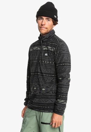 Schwarzer Fleece-Pullover mit geometrischen Mustern in Grau und Grün. Hat einen hohen Kragen und einen Reißverschluss vorne. Kombiniert mit grünen Cargohosen.