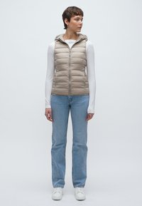 Μπεζ αμάνικο μπουφάν με κουκούλα, με κάθετο καπιτονέ σχέδιο, εμπρόσθιο φερμουάρ, φορεμένο πάνω από λευκό πουκάμισο με μακριά μανίκια και συνδυασμένο με μπλε τζιν.